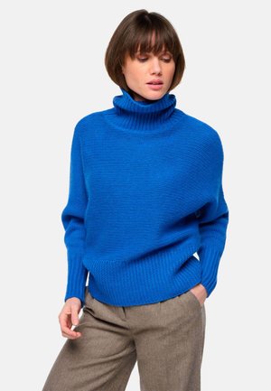 Pull en maille bleu à col roulé avec une texture côtelée et une coupe décontractée, porté avec un pantalon à motifs marron.