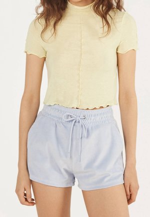 Femme portant un haut court à manches courtes jaune clair et un short bleu clair à taille élastique et cordon de serrage, debout avec les bras détendus.