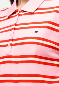 Πόλο μπλούζα με οριζόντιες ρίγες σε ροζ, κόκκινο και λευκό, με γιακά και μικρό λογότυπο στο αριστερό μέρος του στήθους.