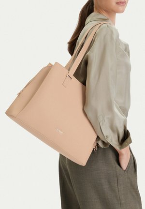 Žena v světle olivové blůze a šedých kalhotách nese přes rameno velkou béžovou koženou tašku Lasocki.