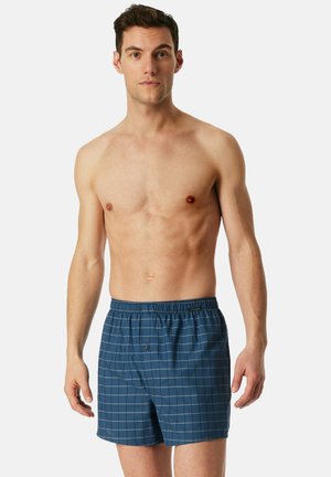 Uomo senza maglietta che indossa boxer blu a quadretti, in piedi contro uno sfondo chiaro e uniforme, rivolto in avanti con un'espressione neutra.