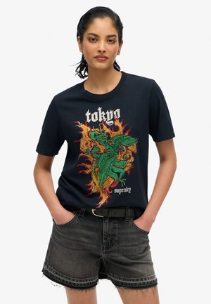 T-shirt in cotone blu scuro con una grande grafica di un drago colorato e il testo "tokyo". Abbinato a pantaloni shorts in denim neri sbiaditi e una cintura nera.
