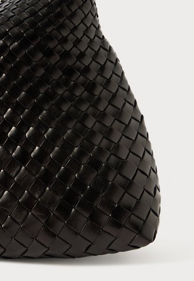 Mustan kudotun nahkakäsilaukun teksturoidulla pinnalla, jossa on ainulaatuinen kolmiomainen muoto ja saumaton design. Ei näkyvää metalliosaa.