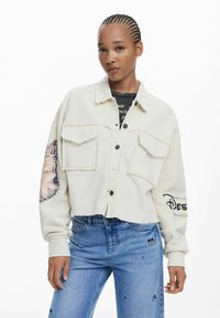 Giacca corta in corduroy bianca con due tasche sul petto, orlo sfrangiato e design grafico sulla manica. Abbinata a jeans azzurri chiari con piccoli motivi.