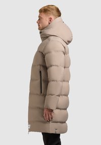 Beige pufferjakke med hætte, der har et quiltet design, sidelomme med lynlås og en lang pasform, der når til midt på lårene. Glat tekstur.
