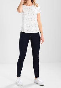 Camisa blanca de manga corta con pequeños patrones de anclas, combinada con jeans ajustados oscuros y zapatillas blancas, de pie sobre un fondo gris.