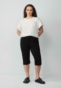 Witte katoenen t-shirt met een kleine bloemenborduursel op de linkerborst, gecombineerd met zwarte knielange broek en zwarte instap schoenen.