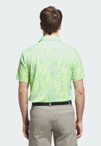 Koszulka polo w jasnej zieleni z gradientowym wzorem, krótkie rękawy i kołnierzyk. Noszona z beżowymi spodniami i czarnym paskiem. Widok od tyłu.