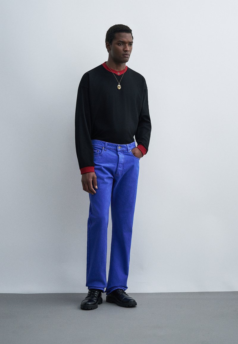 Homme debout contre un mur gris, portant un pull noir sur une chemise rouge, un jean bleu, des chaussures noires et un collier avec un pendentif en or.