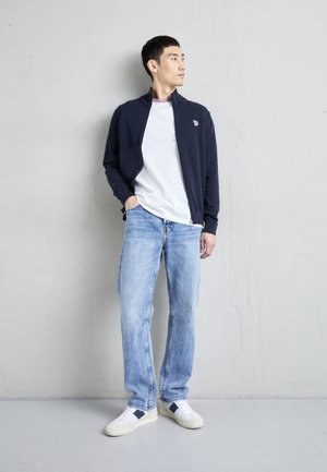 Tumesinine tõmblukuga jope valge T-särgi kohal, koos kulunud siniste teksade ja siniste aktsentidega valgete spordialustega. Sujuvad tekstuurid, lõdva istuvusega.