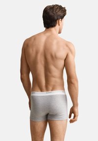 Homme debout, tourné de dos, portant un boxer gris avec une ceinture blanche marquée "GANT", sur un fond clair uni.