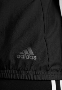 Tissu noir Adidas avec ourlet cousu et logo Adidas en gris foncé au-dessus de deux rayures blanches verticales sur fond noir.