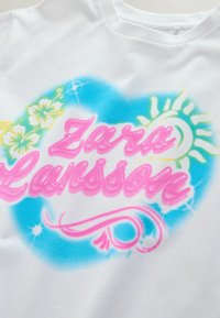 Chemise blanche avec un motif coloré à l'aérographe présentant "Zara Larsson" en rose, un fond de cœur bleu, des fleurs d'hibiscus jaunes, un soleil et des volutes décoratives.