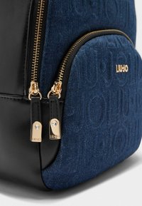 Zwarte en donkerblauwe rugzak met een denimtextuur, een reliëflogo en ritsen in goudkleurige toon met decoratieve hardwareaccenten.