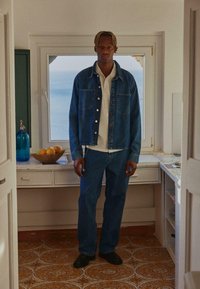 Komplet od traper jakne i hlača u tamnoplavoj boji, uparen s bijelom košuljom s ovratnikom. Glatka tekstura i klasičan kroj. Crne cipele zaokružuju izgled.