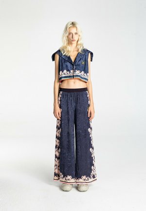 Persona alta con cabello rubio largo vistiendo pantalones anchos de pierna con estampado floral azul marino y chaqueta corta sin mangas a juego, de pie sobre fondo blanco.