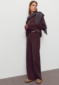 Bruin geribbeld loungewear set, losse broek en trui, met een grijze sjaal over de schouders gedrapeerd. Beige instapschoenen maken de look af.