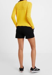 Žena v žlutém pleteném svetru s dlouhým rukávem, černých šortkách a černých teniskách, stojí s rukama v kapsách, otočena zády.