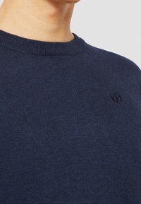 Námorně modrý pletený svetr s kulatým výstřihem a jemnou strukturou, se malým logem na hrudi. Hladká tkanina.