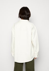 Persona con cabello corto y oscuro, vistiendo una camisa blanca de manga larga oversize y pantalones verdes oliva, frente a un fondo claro y liso.