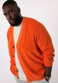 Orange gestrickte Strickjacke mit V-Ausschnitt, Knopfleiste und zwei Vorderseiten-Taschen. Mit gerippter Textur und kontrastierenden dunklen Knöpfen.