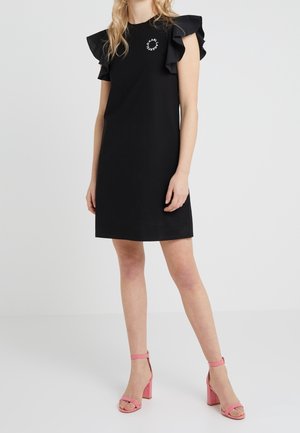 Schwarzes kurzes Kleid mit Rüschensäumen und einem kreisförmigen grafischen Detail. Kombiniert mit rosa Riemchenheels und einer glatten Textur.