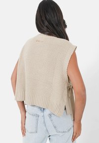 Gilet en maille beige avec des manches courtes, motif texturé, ourlet côtelé et un détail de nouage à l'arrière. Porté sur un jean bleu clair.
