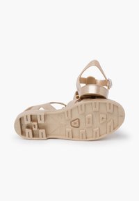 Beige rubber sandal with a textured sole, featuring an adjustable strap and a circular accent. Tread design consists of square and rectangular patterns.