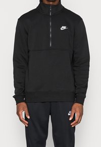 Svart Nike-tröja med hög krage och design med halvdragkedja. Mjuk, texturerad tyg med en vit logotyp på vänster bröst. Enkelt, sportigt utförande.