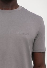 Grå t-skjorte laget av en bomullsblanding. Den har klassisk rund hals, korte ermer og et preget logo "ARMANI EXCHANGE MILANO/NEW YORK" på brystet.
