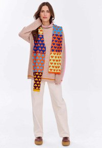 Strickpullover in sanftem Pink mit hohem Kragen, kombiniert mit einem bunten Schal mit Herzmustern in Blau, Orange, Gelb und Lila. Cremefarbene Hosen.