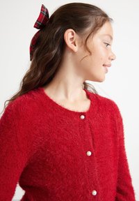 Rote, fluffige Strickjacke mit rundem Ausschnitt und drei Perlenknöpfen an der Vorderseite. Kombiniert mit einer karierten Haarschleife.
