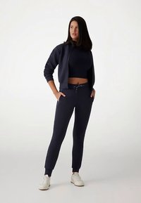 Felpa con cappuccio blu navy con zip abbinata a pantaloni jogging aderenti. Tessuto morbido, vita elastica con coulisse e tasche laterali. Scarpe da ginnastica bianche.