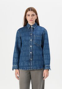 En ung kvinne med langt, brunt hår har på seg en mønstret denimskjorte og grå bukser, og står mot en enkel hvit bakgrunn.