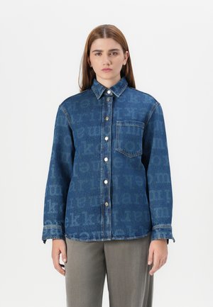 A young woman with long, brown hair wears a patterned denim shirt and gray trousers, standing against a plain white background.