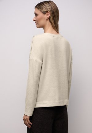 Top beige chiaro a maniche lunghe con vestibilità ampia, tessuto morbido e scollo tondo, caratterizzato da bottoni coordinati sulla spalla.