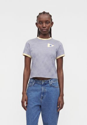 Junge Frau mit geflochtenem Haar, die ein hellblau-weiß gestreiftes T-Shirt mit gelbem Abschluss und blaue High-Waist-Jeans trägt, vor weißem Hintergrund steht.