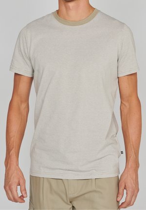 Man draagt een lichtbeige dun gestreept T-shirt met korte mouwen en een kaki broek, staand tegen een effen achtergrond.