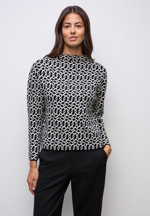 Black and white patterned top with a high neckline, geometric design, and long sleeves, paired with dark trousers. Smooth texture, fitted style.