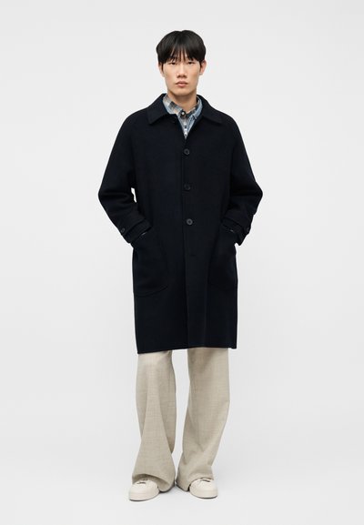 Νεαρός άνδρας στέκεται φορώντας ένα μαύρο παλτό με κουμπιά πάνω από ένα καρό πουκάμισο, μπεζ παντελόνι με φαρδιά πόδια και λευκά αθλητικά παπούτσια, μπροστά από ένα απλό φόντο.