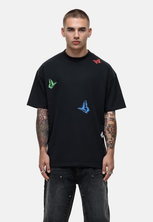 Black cotton t-shirt featuring green, red, and blue butterfly prints. Short sleeves, loose fit, and side slits at the hem.