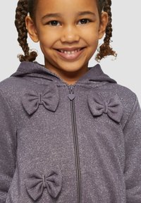 Lila Zip-Hoodie mit funkelnder Textur, versehen mit drei Schleifen auf der Vorderseite und einer Kapuze. Das Material wirkt weich und bequem.