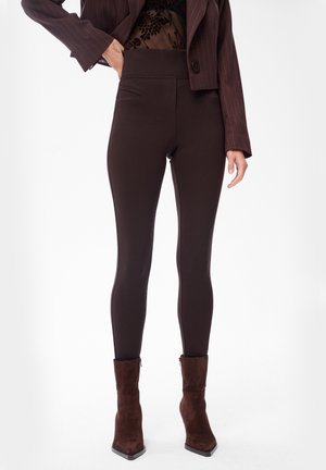 Frau trägt dunkelbraune, eng anliegende Hose, passende Wildleder-Stiefeletten, transparente schwarze Bluse mit floralem Muster und einen gekürzten Nadelstreifen-Blazer, steht mit der Hand in der Hüfte.