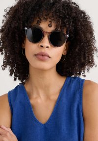 Runde Sonnenbrillen mit schwarzen Linsen, Fassung im Schildpatt-Muster, getragen von einer Person mit lockigem Haar und einem blauen ärmellosen Oberteil.