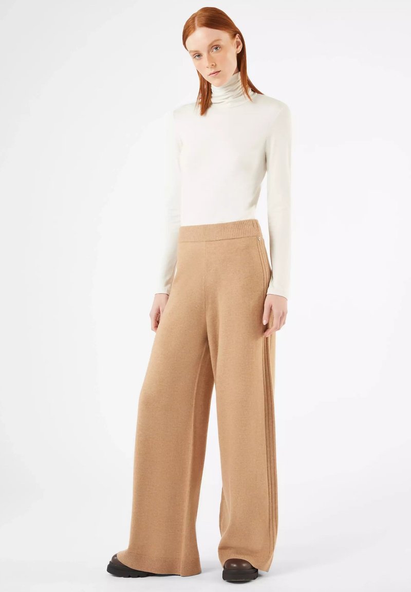 Beige weite Strickhosen mit einem gerippten Bund, kombiniert mit einem figurbetonten weißen Rollkragenpullover. Glatte Textur, leicht ausgestellte Silhouette.