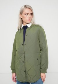 Olīvzaļš pārvilkts jakas ar gludu apdari, pogām ar spiedpogu, sānu kabatām un brīvu piegriezumu. V-veida kakla izgriezums ar baltu kreklu zem tā.