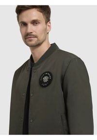 Tume roheline bomber-jakk ümmarguse musta plaastriga, millel on valge tekst, nööbid ja soonikkoes krae. Sile tekstuur, igapäevane disain.