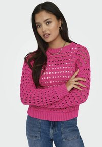 Roze gehaakte trui met open weefdesign, geribbelde manchetten en zoom, gedragen over blauwe jeans. Heeft een ronde hals en een losse pasvorm.
