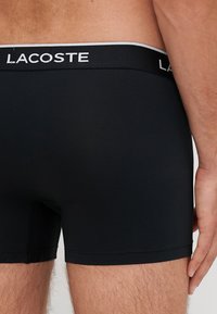 Μαύρα εσώρουχα boxer με λωρίδα ελαστική και λευκό λογότυπο Lacoste στη ζώνη. Διαθέτουν στενή εφαρμογή και λεία υφή. Χωρίς ορατά σχέδια.