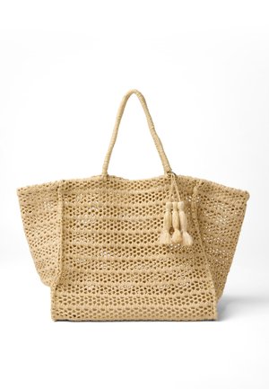 Geflochtener beiger Tragetasche mit offenem Strickmuster, zwei Griffen und dekorativen Quasten, die an einer Seite hängen, vor weißem Hintergrund dargestellt.